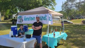 Two people stand under a tent labeled "Hometown Grounds" at an outdoor event. The table displays syrups, cups, and other coffee supplies. Trees and parked cars are visible in the background on a sunny day.