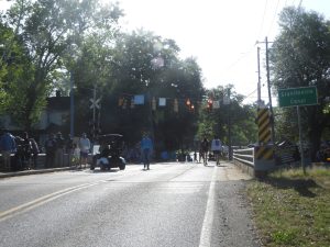 A road leading to a bridge is seen in daylight. People walking and a golf cart are visible on the street. There is a sign that reads "Graniteville Canal" on the right. Traffic lights hang overhead, and trees line the road.