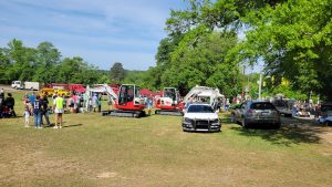 A community event with people gathered around multiple small excavators on display, surrounded by greenery. A black car and another vehicle are parked on the grass, and a fire truck is visible in the background.