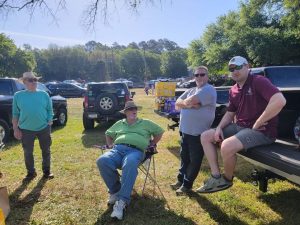 Four men are gathered outdoors near parked trucks. One is sitting in a camping chair, while the others stand nearby. They are wearing casual clothing, and the setting appears to be a grassy area with trees and other vehicles in the background.