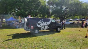 A black van with "105.7 The Bull" logo is parked on a grassy area. People are under tents in the background, and there are trees and parked cars around. It's a sunny day in a park-like setting.