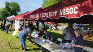 People are gathered under a red tent labeled "Grantville Cemetery Assoc." They are seated and standing near a table with papers and a bell. Several other tents and a fire truck are visible in the background on a sunny day.