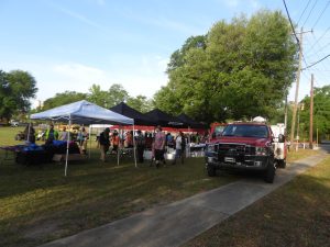 A community event with people gathered under several tents. A red and white vehicle is parked nearby on a grassy area. Tables with items are set up under the tents. Trees and a clear blue sky are in the background.