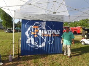 A man stands under a white canopy next to a large blue banner with orange and white text reading "Graniteville Fishing Rodeo." A fish illustration is on the banner. A red fire truck is visible in the background on a grassy area.