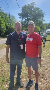 Two men standing outdoors, posing for a photo. One is wearing a navy shirt with a badge and jeans, the other a red shirt, shorts, and a cap. Both are smiling, with tents and people in the background on a sunny day.