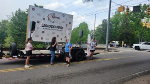 Four people are on a street next to a Bridgestone stage on a trailer. Two are handing microphones to a participant holding a stick, while another stands nearby. Traffic lights and trees are in the background.
