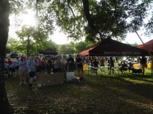 A group of people gathers under and around tents labeled "Granteville Cemetery Assoc" in a grassy, tree-shaded area. Chairs and tables are set up, with some people sitting and others standing. Sunlight filters through the trees.