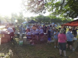 A large group of people gathers outdoors under trees during a daytime event. They are working at tables with supplies and boxes. Some tents are visible in the background. Sunlight filters through the leaves, creating dappled light on the ground.