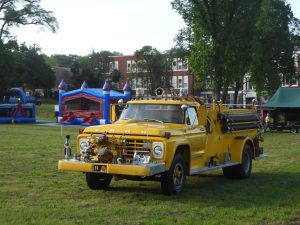A vintage yellow fire truck is parked on a grassy field with bouncy houses and trees in the background. Several buildings are visible under a clear sky, and the scene has a festive, community gathering atmosphere.