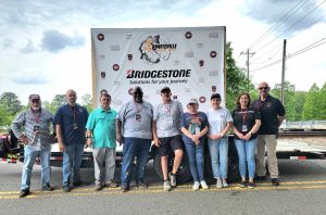 A group of nine people standing in front of a backdrop displaying "Bridgestone" and other logos. They are outdoors, possibly at an event. The backdrop includes the text "Solutions for your journey." Trees and power lines are visible in the background.