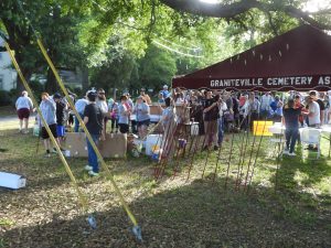 A large group of people gather outdoors near a tent labeled "Graniteville Cemetery Association." Fishing rods are leaning against the tent. The scene is shaded by trees, with tables set up and boxes scattered around.