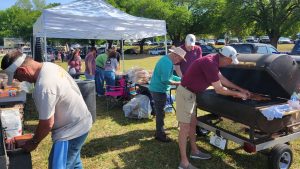 A group of people are gathered outdoors for a barbecue. Two men are tending to a large grill, while others prepare food nearby. There is a white canopy tent, chairs, and parked cars in the background, with trees providing shade.