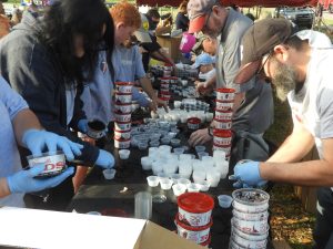 A group of people wearing gloves work at a long table outdoors, organizing and filling cups with soil from containers labeled "DS". They are participating in a community activity on a sunny day.