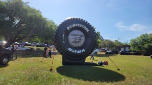 A large inflatable Bridgestone tire is displayed outdoors in a grassy area. Several people stand around, and there are trees and vehicles in the background under a clear blue sky.