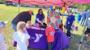Children and adults gather at a YMCA booth under a pink canopy in a park. A woman stands behind a table with a colorful prize wheel. A child in a red shirt waits, while others watch and a man fills out a form.