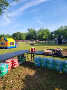 A grassy field with tables holding stacks of soft drink packs, water bottles, and condiments. In the background, there's a colorful inflatable bounce house and booths visible with groups of people scattered across the scene.