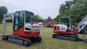 Two red and white excavators on display in a grassy area. The one on the left is a 12R/CW, and the one on the right is a TB370. Tents and people in the background under a partly cloudy sky.