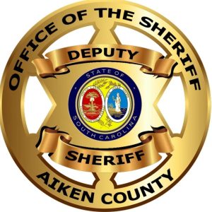 Gold badge with "Office of the Sheriff Aiken County" and "Deputy Sheriff." Center features South Carolina state seal with palmetto tree and figures. Circular design with text on ribbon banners.