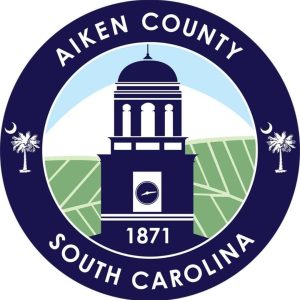A circular seal for Aiken County, South Carolina, featuring a central clock tower with "1871" below. Surrounding text reads "Aiken County South Carolina." Crescent moons and palmetto trees are on either side against a backdrop of hills.